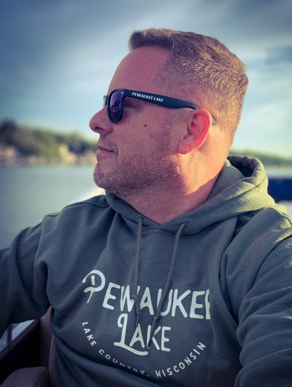 Man on a pontoon bat, looking to the side showcasing his Pewaukee Lake sunglasses and grey hoodie that says "Pewaukee Lake, Lake Country, Wisconsin"