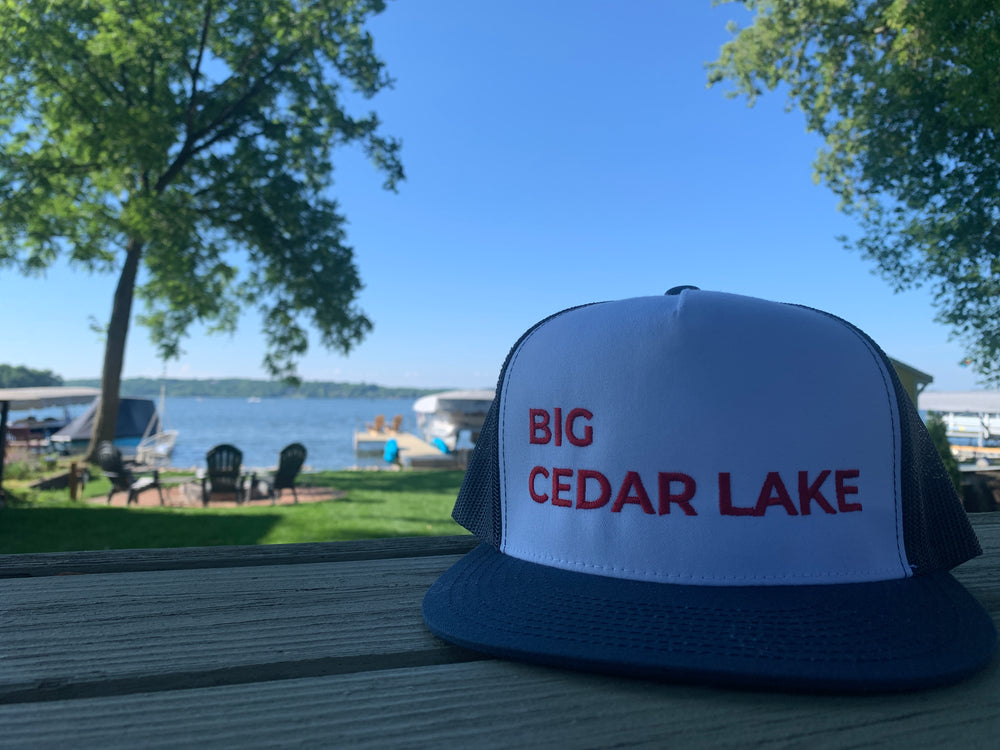 Hat with white front and "Big Cedar Lake" written in large text in all caps, resting on a picnic table with a grassy area with firepit and chairs as well as pier, pontoon boat docked and Big Cedar Lake in Wisconsin in the background