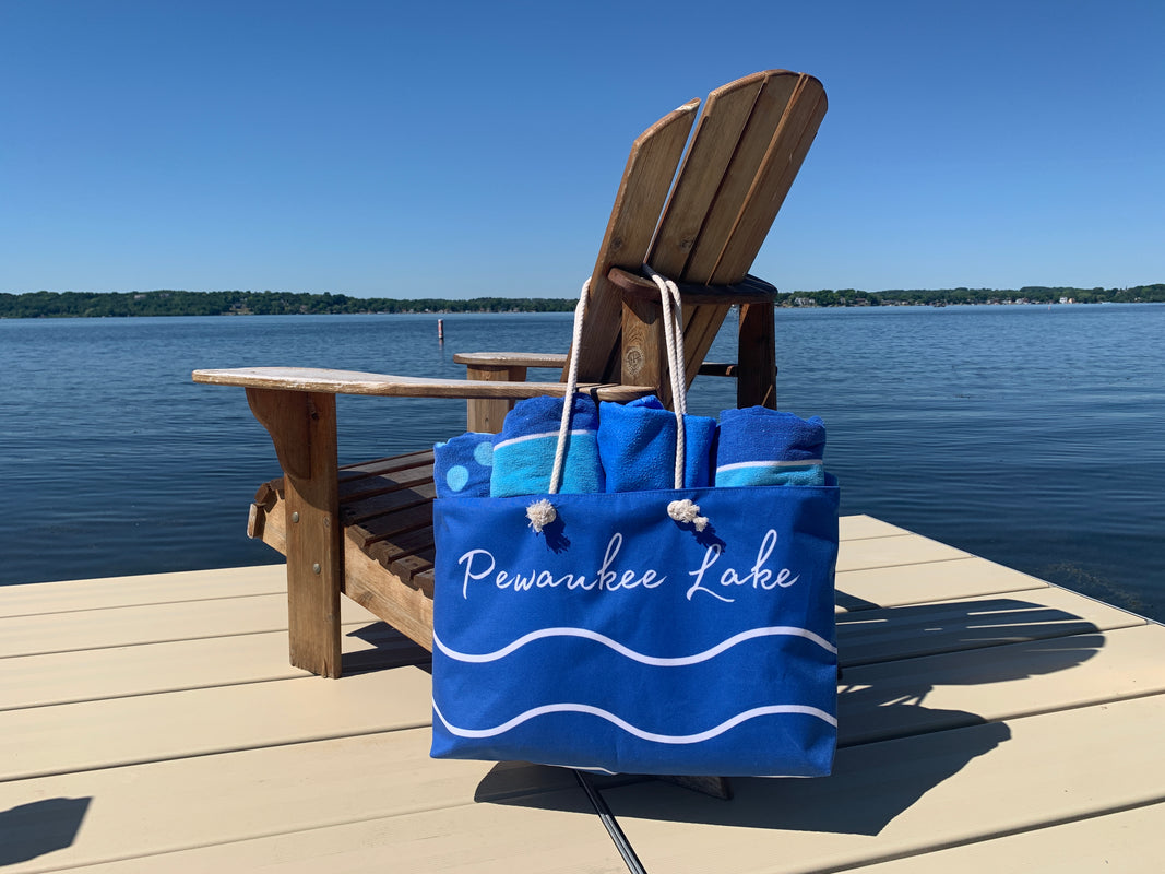 Blue tote bag with white wavy lines and the words "Pewaukee Lake" holding 4 blue rolled up towels hanging on an Adirondack chair on a pier overlooking Pewaukee Lake in Wisconsin