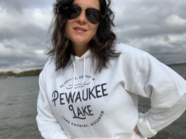 Woman posing in front of Pewaukee Lake, WI wearing a white hoodie sweatshirt that says "Pewaukee Lake, Lake Country, Wisconsin" in black font with the coordinates above it