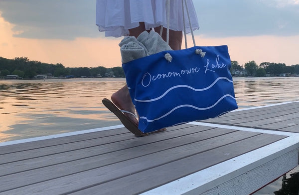 Closeup of a pretty blue beach tote bag that says "Oconomowoc Lake" in white script with 2 wavy lines below it, person holding it is walking on the pier overlooking Oconomowoc Lake in Wisconsin at sunset. The bag is being sold online by Lake Country Vibes