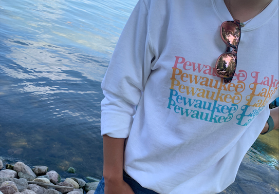 White crew neck sweatshirt with "Pewaukee Lake" written in fun font 5 times stacked in a different bright color, person wearing it you cannot see their face but you can see they are standing on rocks in front of Pewaukee Lake in Wisconsin