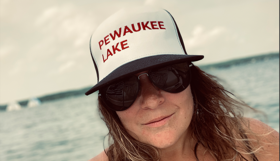 Woman smiling, wearing a "Pewaukee Lake" trucker hat with Pewaukee Lake in Wisconsin behind her. Hat sold by Lake Country Vibes online shop